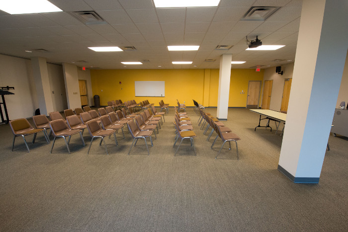 Large room with lots of chairs front table, media cart and ceiling mounted projector.