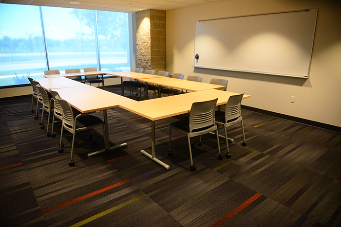 Room with wall of windows, dry erase board and tables/chairs arranged in large rectangle.